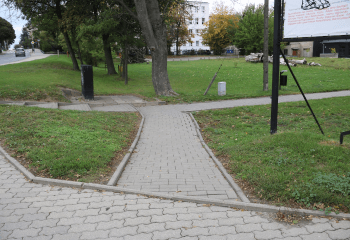 Zdjęcie. Na pierwszym planie chodnik z betonowej kostki z małym uskokiem. W tle trawnik.
