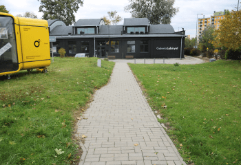 Zdjęcie. Na pierwszym planie chodnik z betonowej kostki, prowadzący do budynku galerii. Po lewej stronie widać fragment żółtego kiosku. Po obu stronach chodnika trawnik.